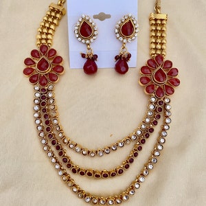 Puede incluir: Un collar de oro con tres hileras de piedras rojas y transparentes, y un par de pendientes a juego con piedras rojas y transparentes. El collar tiene un dise&ntilde;o floral con piedras rojas en el centro.