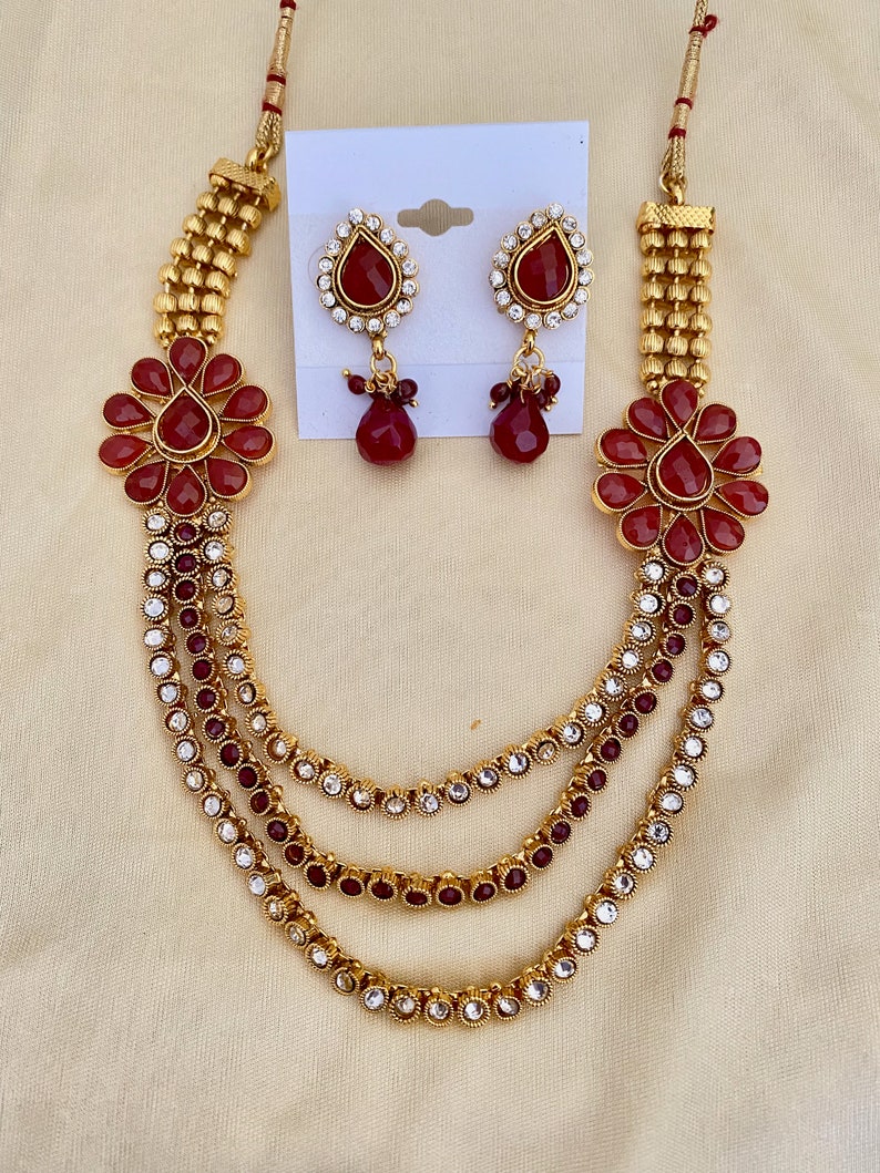 Puede incluir: Un collar de oro con tres hileras de piedras rojas y transparentes, y un par de pendientes a juego con piedras rojas y transparentes. El collar tiene un dise&ntilde;o floral con piedras rojas en el centro.