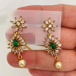 May include: A pair of gold-toned earrings featuring a floral design with clear and emerald-green gemstones. Each earring has a pearl drop. The earrings are displayed on a white background.