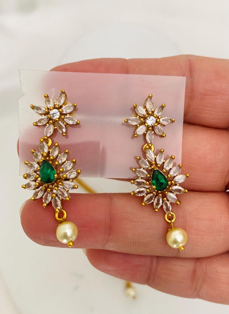 May include: A pair of gold-toned earrings featuring a floral design with clear and emerald-green gemstones. Each earring has a pearl drop. The earrings are displayed on a white background.