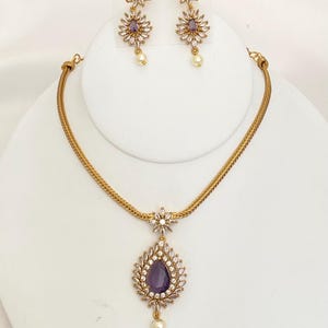 May include: A gold-toned jewelry set featuring a necklace and matching earrings. The necklace has a teardrop pendant with a purple center stone, surrounded by clear crystals and a dangling pearl. The earrings mirror the pendant design, with a floral motif.