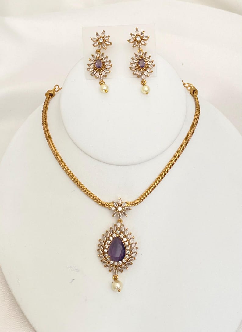 May include: A gold-toned jewelry set featuring a necklace and matching earrings. The necklace has a teardrop pendant with a purple center stone, surrounded by clear crystals and a dangling pearl. The earrings mirror the pendant design, with a floral motif.
