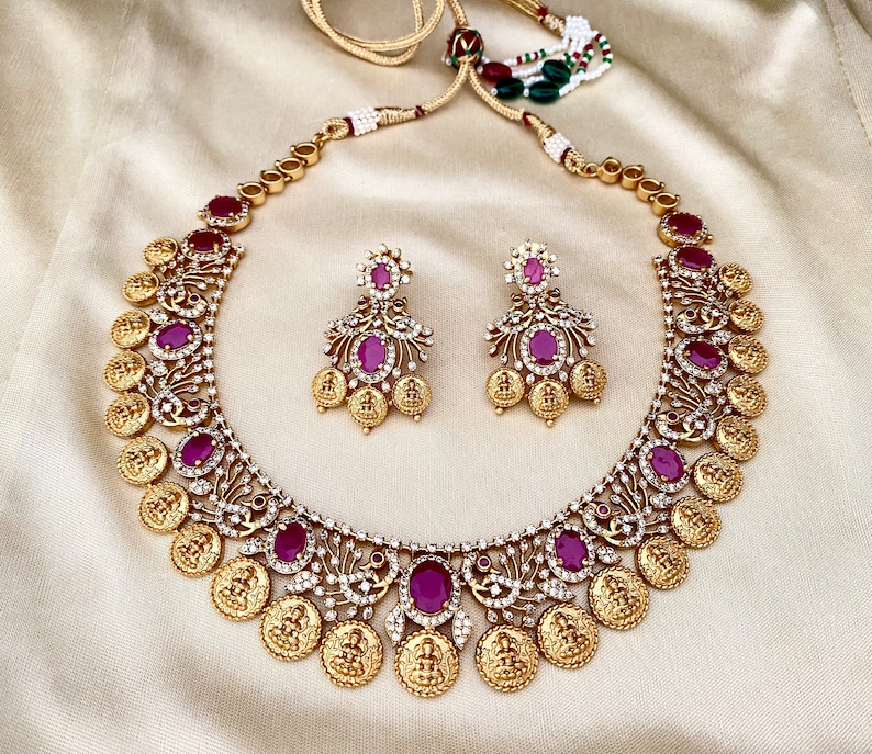 May include: A gold necklace with a coin design and pink gemstones. The necklace is paired with matching earrings.