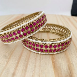 May include: Two gold bangles with pink stones and white pearl accents.
