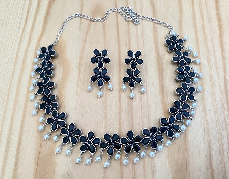 Op de afbeelding: Een zilveren ketting met zwarte bloembedels en witte parel accenten. Passende oorbellen met zwarte bloembedels en witte parels.