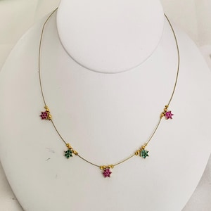 May include: A delicate gold-tone necklace featuring small, colorful floral accents. The necklace has a thin chain with evenly spaced clusters of red and green gemstones, and small gold beads. The necklace is displayed on a white bust.