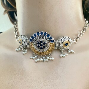 May include: A silver and gold choker necklace with a large blue and gold centrepiece. The centrepiece is surrounded by smaller blue stones and has a floral design. The necklace has a chain with small white pearl drops.