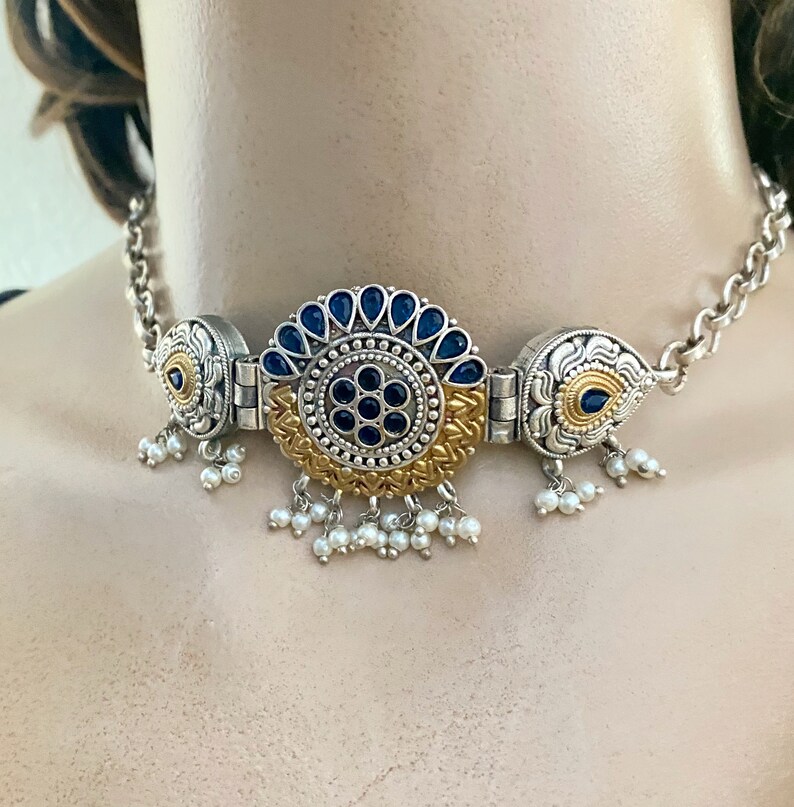 May include: A silver and gold choker necklace with a large blue and gold centrepiece. The centrepiece is surrounded by smaller blue stones and has a floral design. The necklace has a chain with small white pearl drops.