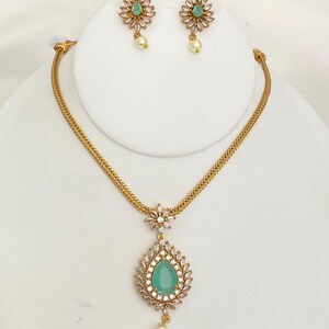 May include: A gold-toned jewelry set featuring a necklace and earrings. The necklace has a teardrop-shaped pendant with a teal center stone, surrounded by clear crystals, and a pearl drop. The earrings mirror the pendant design with a flower-shaped top and pearl drop.
