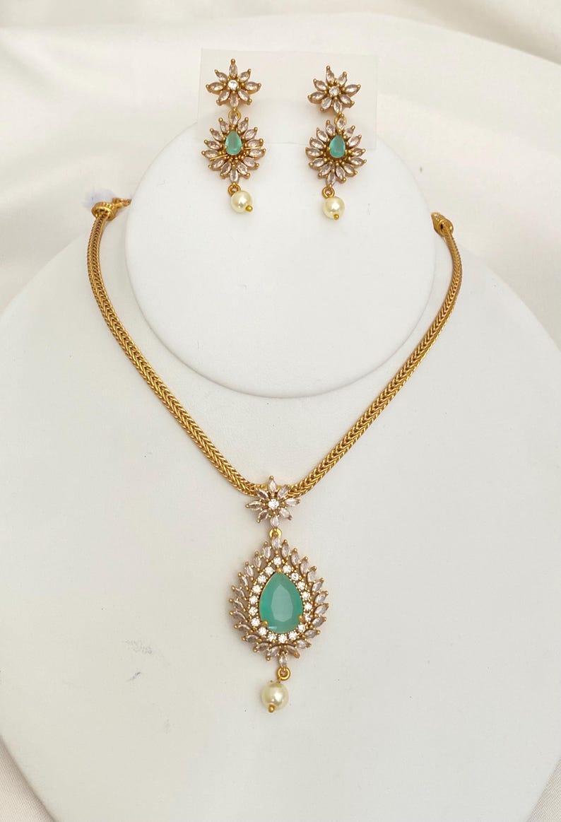 May include: A gold-toned jewelry set featuring a necklace and earrings. The necklace has a teardrop-shaped pendant with a teal center stone, surrounded by clear crystals, and a pearl drop. The earrings mirror the pendant design with a flower-shaped top and pearl drop.