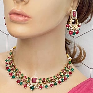 May include: A gold necklace with green and red stones and white pearls. The necklace has a square-shaped pendant with a red stone in the center. The necklace is paired with matching earrings with a red stone and green and white stones.
