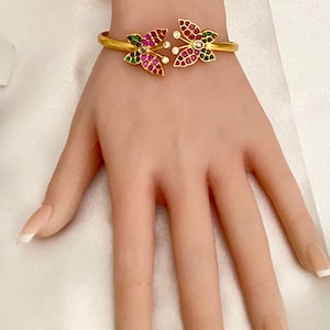 May include: Gold-toned bracelet featuring two butterfly-shaped designs. Each butterfly is adorned with red, green, and white gemstones. The bracelet is open and rests on a hand with manicured nails, showcasing the jewelry's design and color.