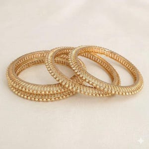 May include: A set of three gold-toned metal bangles featuring a textured surface with a repeating pattern of small raised beads and diagonal etched lines. The circular bracelets are arranged in an overlapping formation, showcasing a metallic finish.