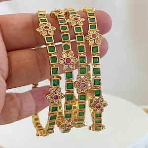 May include: A set of four gold-tone bangles featuring square-cut green stones arranged in a repeating pattern. Each bangle is accented with floral-shaped motifs containing a central round pink stone surrounded by small clear white crystals.