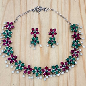 Op de afbeelding: Een zilveren ketting met een bloemenontwerp met afwisselend groene en roze bloemen. De ketting heeft een zilveren ketting en parel accenten. Passende oorbellen met hetzelfde bloemenontwerp zijn inbegrepen.