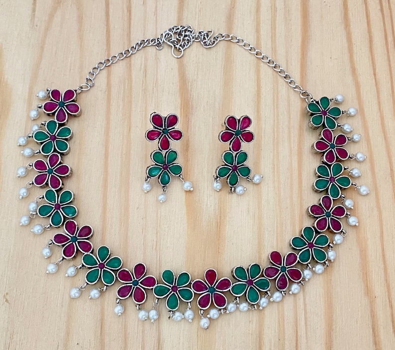 Op de afbeelding: Een zilveren ketting met een bloemenontwerp met afwisselend groene en roze bloemen. De ketting heeft een zilveren ketting en parel accenten. Passende oorbellen met hetzelfde bloemenontwerp zijn inbegrepen.