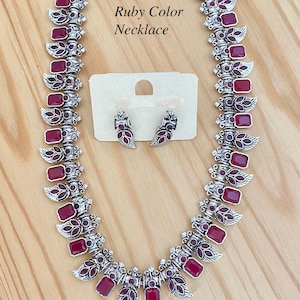 May include: A silver necklace with ruby red stones and matching earrings. The necklace features a design of leaves and the text "Ruby Color Necklace".