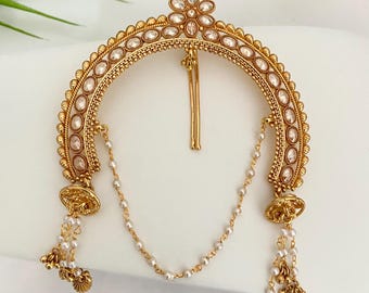 May include: A gold-toned hair accessory with a floral design at the top and a curved, embellished frame. The frame is adorned with clear stones and small pearls, with dangling pearl chains and bell-shaped accents.