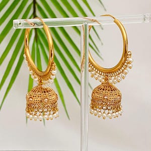May include: A pair of gold-colored hoop earrings with a bell-shaped charm. The charm is intricately designed with a lattice pattern and adorned with small, dangling white pearls. The earrings are displayed on a clear stand against a backdrop of green leaves.