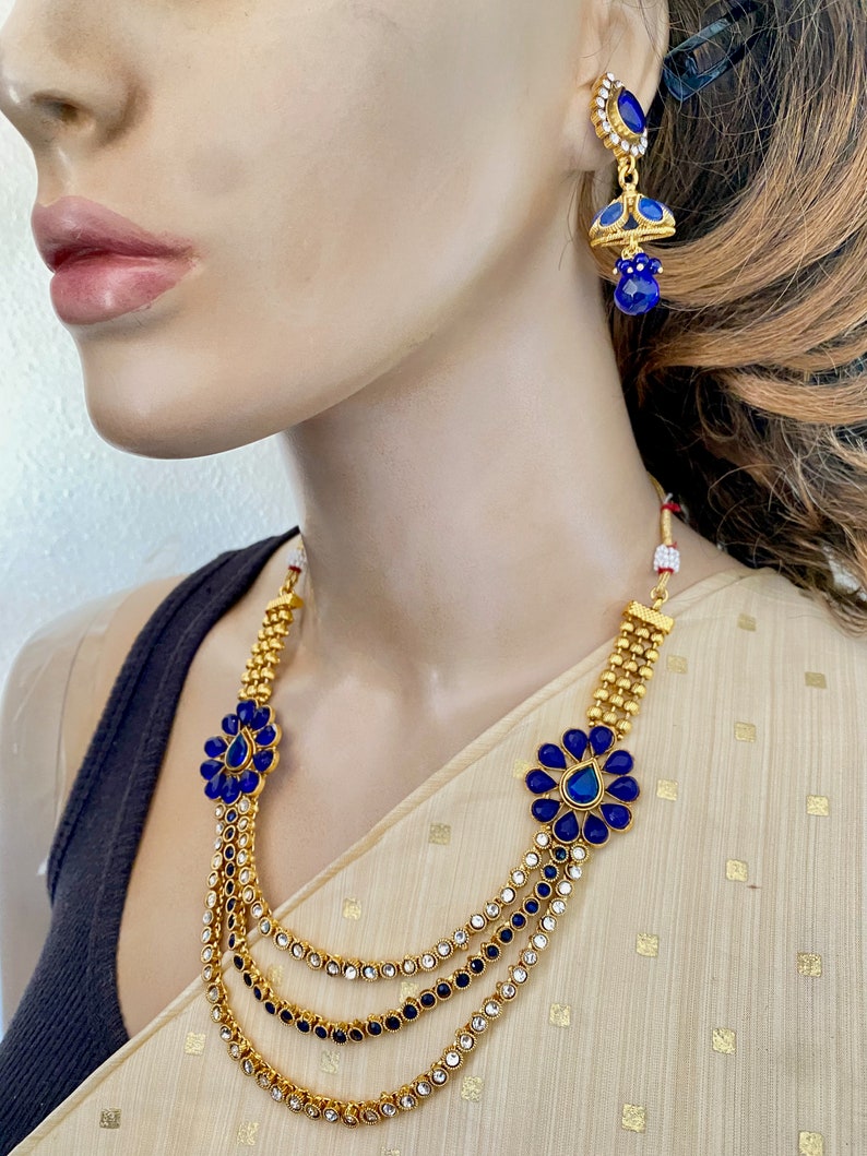 Puede incluir: Un collar de oro con tres hileras de piedras azules y transparentes. El collar tiene un dise&ntilde;o de flor con una piedra azul en el centro. El collar se combina con pendientes a juego con una piedra azul y un colgante.