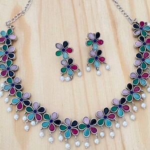 Op de afbeelding: Een zilveren ketting met een bloemenpatroon en parel accenten. De ketting wordt geleverd met bijpassende oorbellen. De bloemen zijn veelkleurig in de kleuren roze, groen, blauw en zwart.