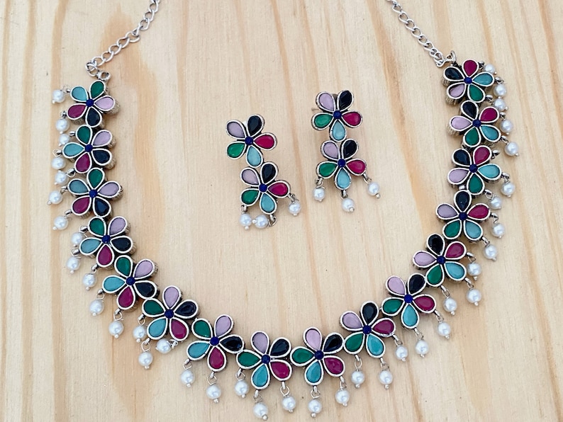 Op de afbeelding: Een zilveren ketting met een bloemenpatroon en parel accenten. De ketting wordt geleverd met bijpassende oorbellen. De bloemen zijn veelkleurig in de kleuren roze, groen, blauw en zwart.