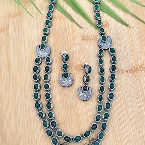May include: A silver necklace and earrings set with green stones. The necklace has two strands of oval-shaped stones and a crescent moon pendant. The earrings are drop earrings with a crescent moon pendant and oval-shaped stones.