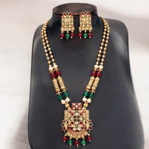 May include: A gold necklace with a square pendant featuring green, red, and white stones. The necklace is adorned with green and red beads. Matching earrings with green and red stones are included.
