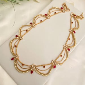 May include: A gold-toned headpiece with multiple strands of delicate chains, adorned with small pearls and vibrant red gemstones. The design features floral accents and is likely intended for special occasions or cultural events.