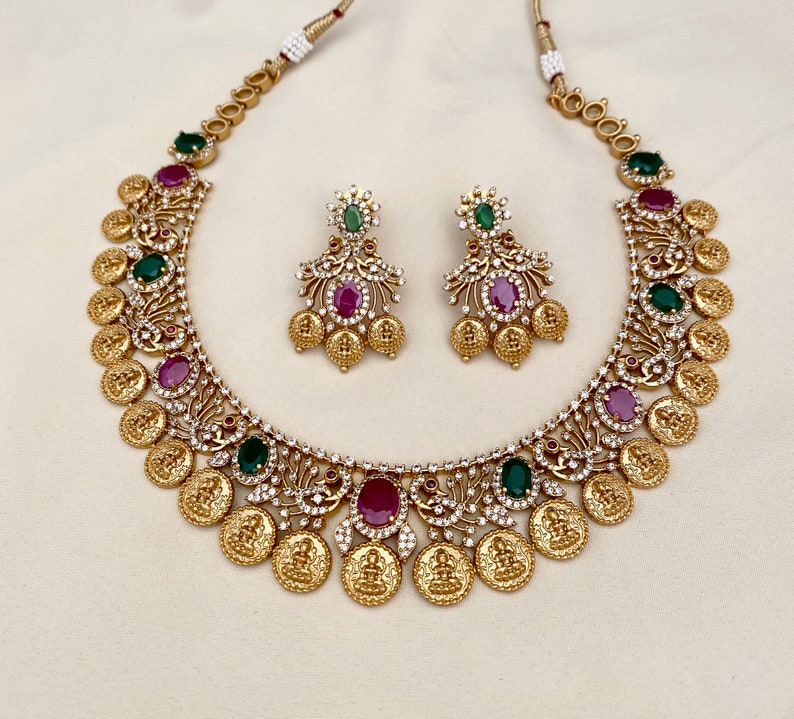 May include: A gold necklace with a coin design and green and pink gemstones. Matching earrings with the same design and gemstones.