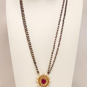 May include: A gold-toned necklace and matching earrings set. The necklace features a double strand of black beads with a central pendant and earrings, each with a red gemstone center surrounded by small white pearls. The jewelry is displayed on a white mannequin.