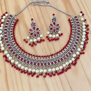 May include: A silver-toned necklace and earring set. The necklace features a series of red teardrop-shaped stones, white pearls, and red beads. The matching earrings have a floral design with red stones and white pearls. The jewelry set is displayed on a wooden surface.