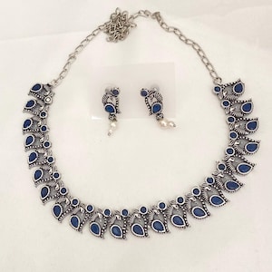 May include: A silver-toned necklace and matching earrings set. The necklace features a chain and a series of ornate, leaf-shaped pendants with dark blue teardrop-shaped stones. The earrings have a similar design, with a small pearl drop.