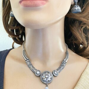 May include: A silver necklace with a large, round pendant featuring a floral design with clear stones. The pendant has a teardrop-shaped stone hanging from it. The necklace is on a mannequin wearing a white top.