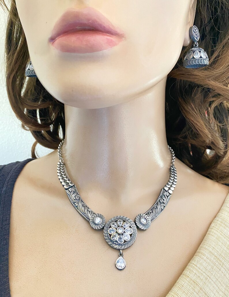 May include: A silver necklace with a large, round pendant featuring a floral design with clear stones. The pendant has a teardrop-shaped stone hanging from it. The necklace is on a mannequin wearing a white top.