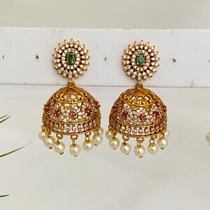 May include: Gold-toned earrings with a floral design. Each earring features a central green stone surrounded by small white stones. The bell-shaped lower portion is adorned with red and white stones and dangling pearls.