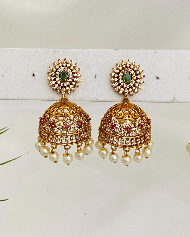 May include: Gold-toned earrings with a floral design. Each earring features a central green stone surrounded by small white stones. The bell-shaped lower portion is adorned with red and white stones and dangling pearls.