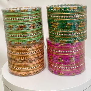 May include: Four stacks of colorful bangles, each wrapped in clear plastic. The bangles are in shades of green, gold, pink, and a darker green. Each bangle has a decorative gold band and small, clear embellishments.