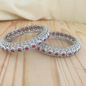 May include: Two silver bangles with intricate designs and pink gemstones. The bangles are stacked on top of each other.