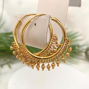 May include: A pair of gold hoop earrings featuring a series of small, textured, spherical charms dangling from the lower half of each hoop. The earrings have a polished finish and a circular design with a thin wire closure mechanism.