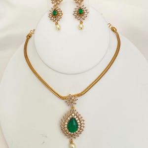 May include: A gold-toned jewelry set featuring a necklace and earrings. The necklace has a teardrop-shaped pendant with a green center stone, surrounded by clear crystals. The earrings and pendant have a floral design with a pearl drop.