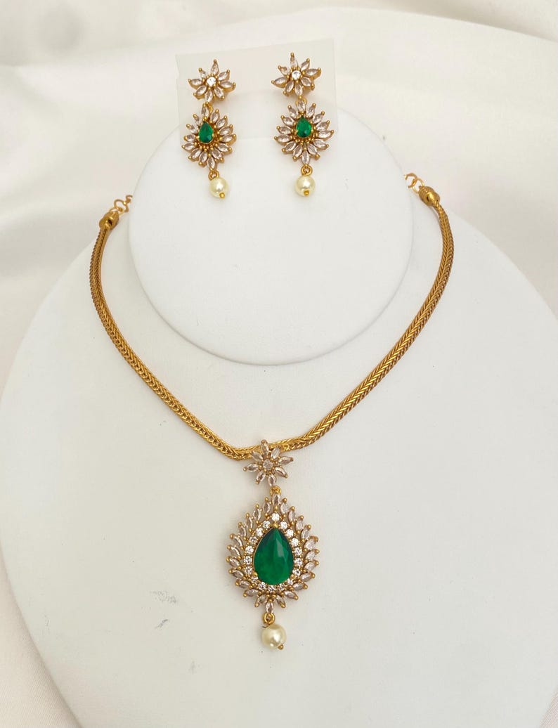 May include: A gold-toned jewelry set featuring a necklace and earrings. The necklace has a teardrop-shaped pendant with a green center stone, surrounded by clear crystals. The earrings and pendant have a floral design with a pearl drop.