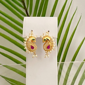 May include: Gold-toned peacock earrings with a red teardrop-shaped gemstone and a green gemstone eye. The earrings are adorned with small white pearls and are displayed on a white stand against a backdrop of green leaves.