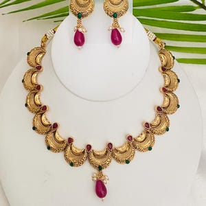 May include: A gold-toned necklace and earring set. The necklace features crescent-shaped pendants with red and green accents. The earrings mirror the design, with a teardrop-shaped, magenta-colored bead and small pearls.
