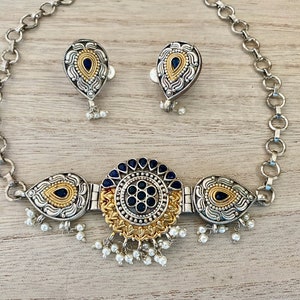 May include: A silver and gold necklace with a round pendant featuring a black and gold design. The pendant is surrounded by three teardrop-shaped pendants with black and gold designs. The necklace is attached to a silver chain. The necklace comes with a pair of matching earrings.