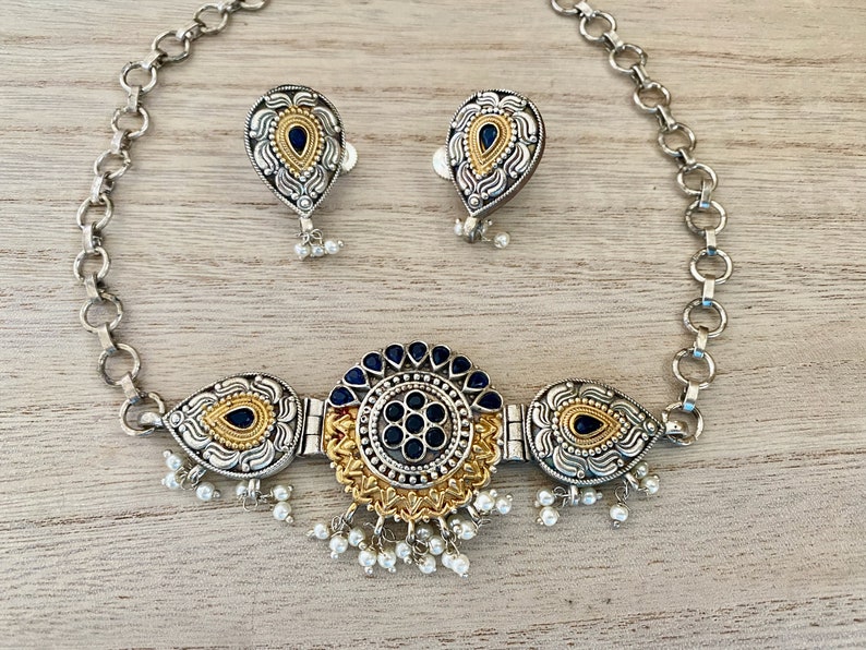 May include: A silver and gold necklace with a round pendant featuring a black and gold design. The pendant is surrounded by three teardrop-shaped pendants with black and gold designs. The necklace is attached to a silver chain. The necklace comes with a pair of matching earrings.