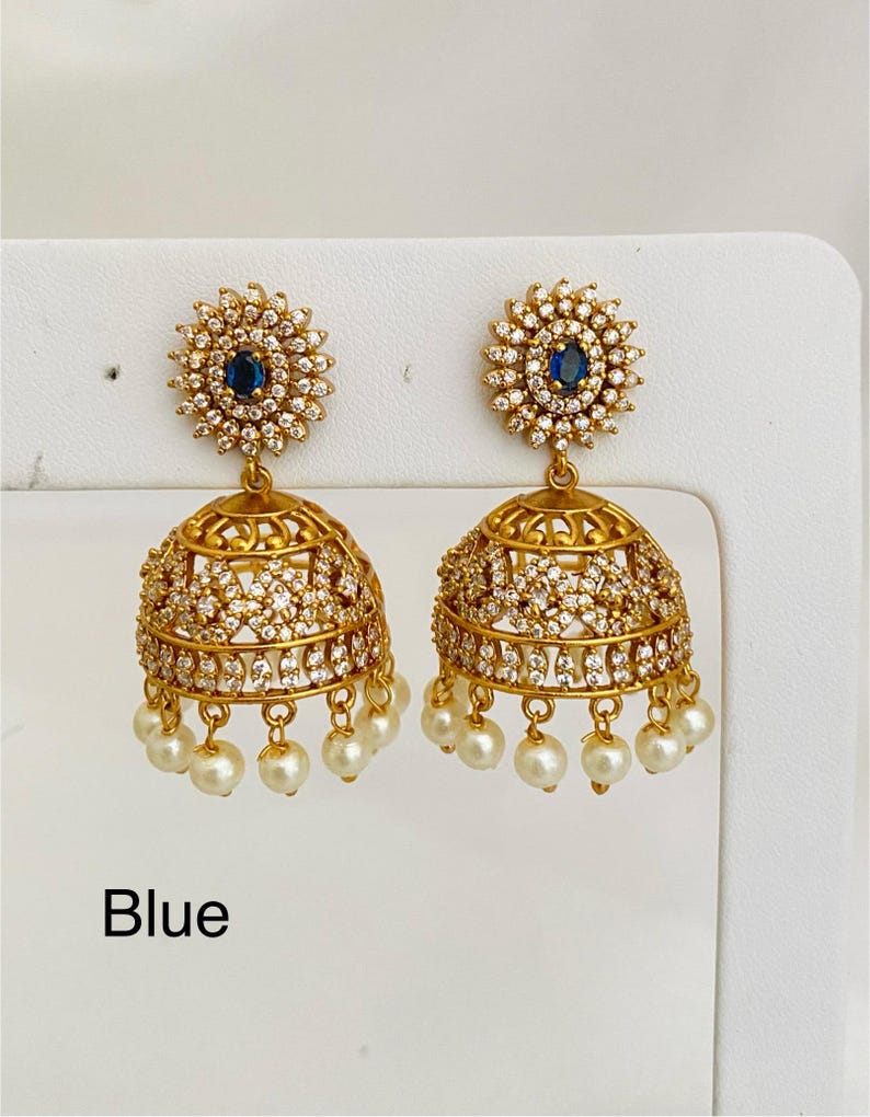 May include: Gold-toned earrings with a floral design and a blue center stone. The earrings have a bell shape, are encrusted with small, clear stones, and feature dangling white pearls. The word "Blue" is printed at the bottom.