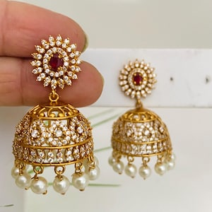 May include: Gold-toned earrings with a floral design, featuring a central red stone surrounded by clear crystals. The bell-shaped earrings are adorned with more crystals and dangling white pearls. The earrings are held up against a white background.