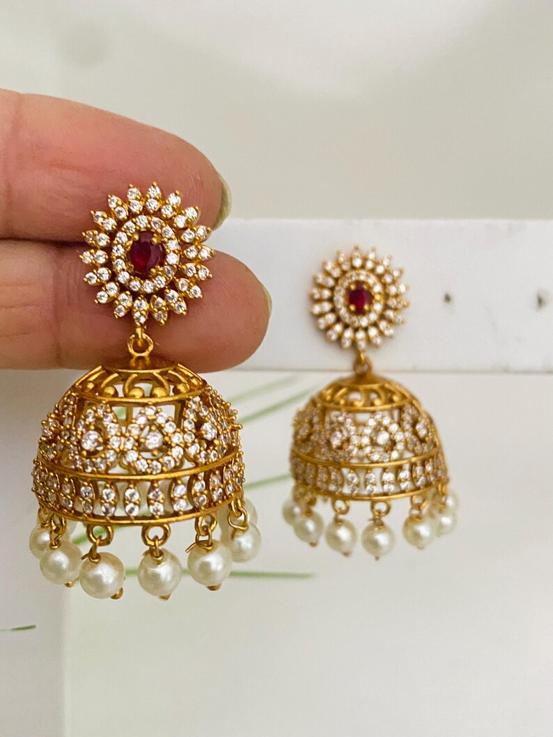 May include: Gold-toned earrings with a floral design, featuring a central red stone surrounded by clear crystals. The bell-shaped earrings are adorned with more crystals and dangling white pearls. The earrings are held up against a white background.