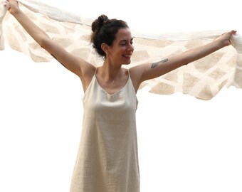 Puede incluir: Una mujer con un vestido lencero color crema sostiene una bufanda estampada. La bufanda tiene un diseño geométrico y se sostiene con los brazos extendidos. El vestido tiene cuello en V.